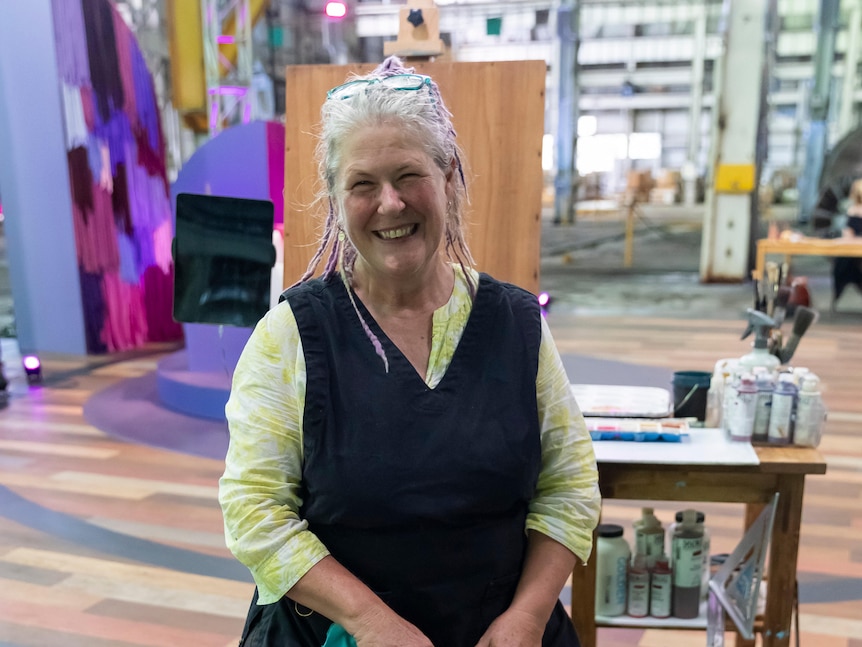A woman with grey and purple dreadlocks, pulled back, smiles at the camera. She sits in front of a table of art supplies