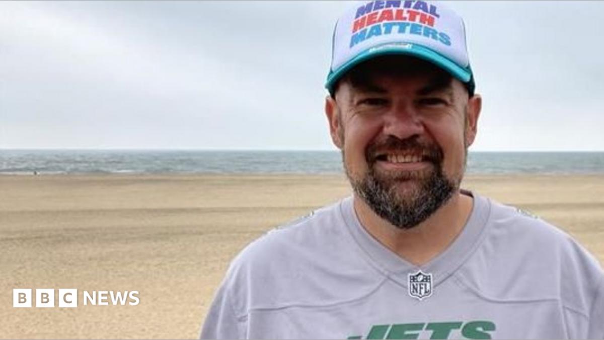 A man standing on a beach smiles at the camera in a head and shoulders image. He has a dark beard and is wearing a white baseball cap with blue peek. Across the front of the cap it says MENTAL HEALTH MATTERS in blue, red and turquoise letters. He is wearing a replica American football top in grey. It has JETS in green capitals across the chest.