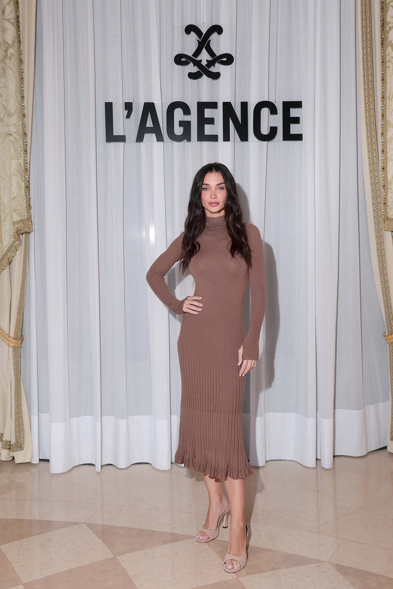 A woman poses in a brown ribbed midi dress in front of the L'AGENCE logo backdrop.