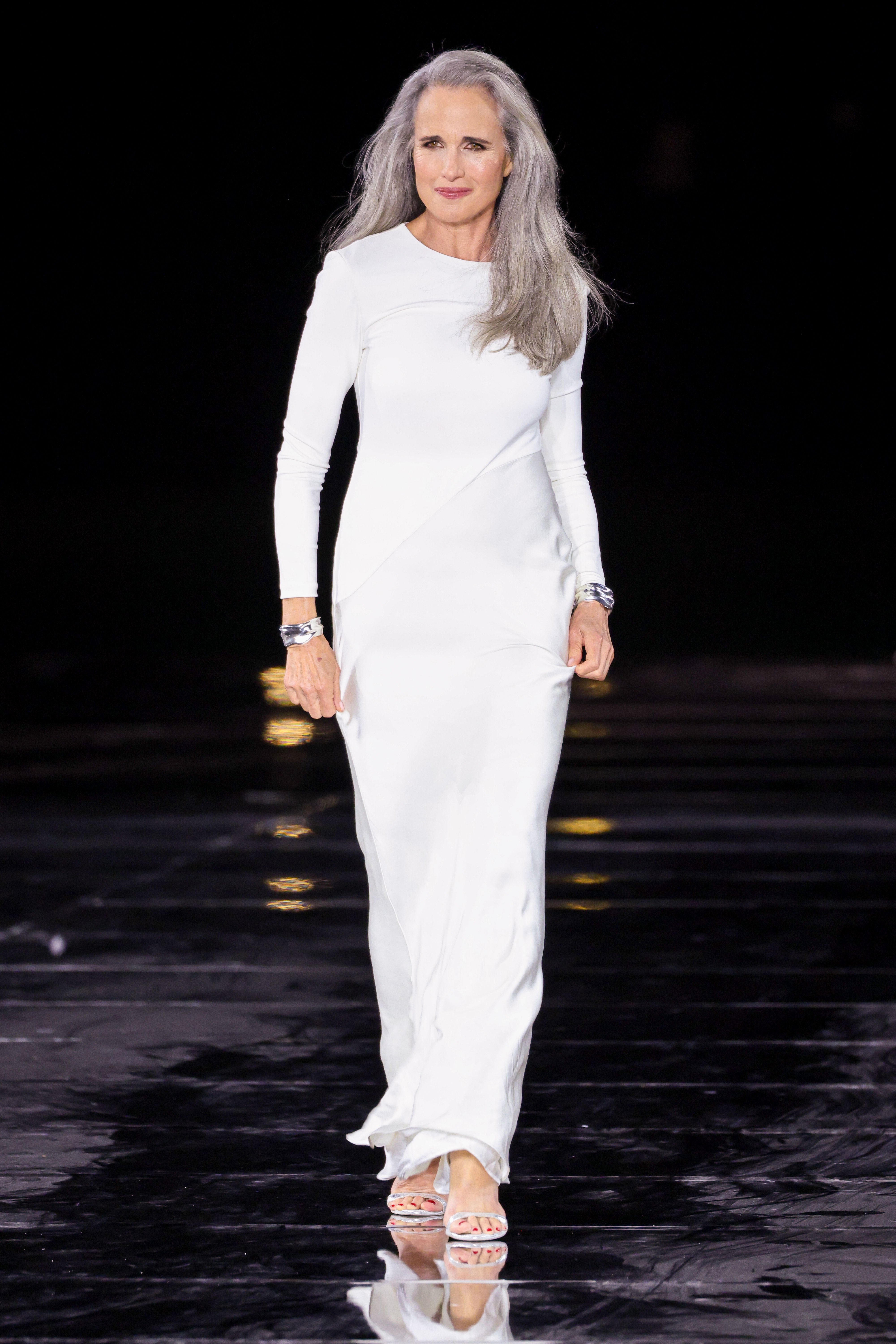 Andie MacDowell on the runway in a long-sleeved white dress with silver bracelets and sandals.