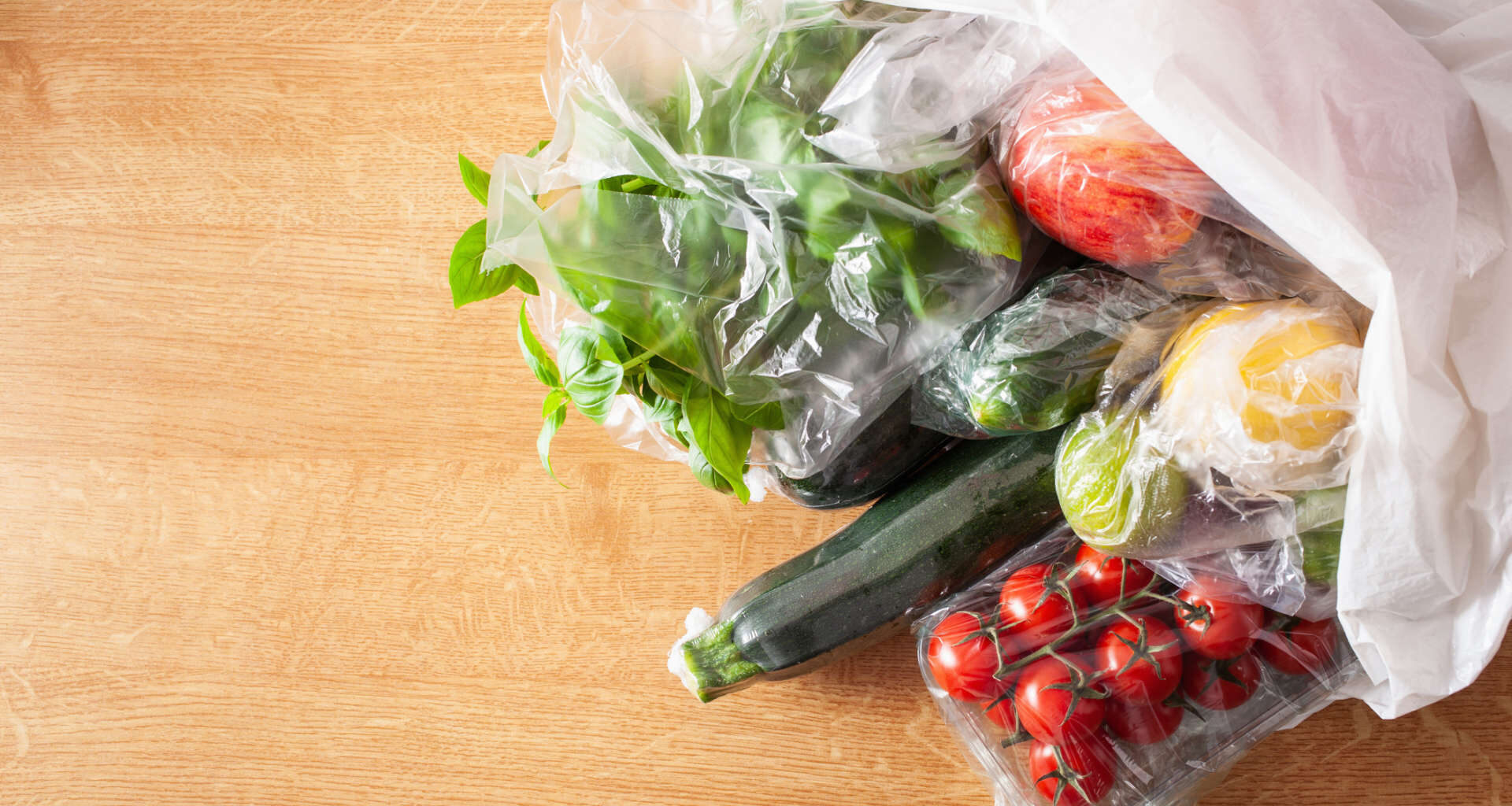 Various fresh vegetables and fruits in plastic bags on a wooden surface.
