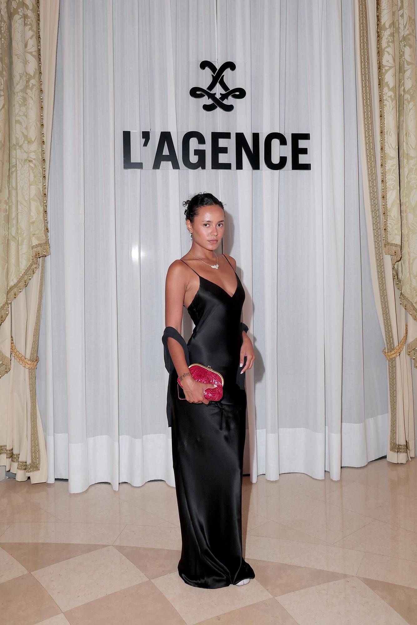 A woman in a black slip dress stands in front of a white curtain with the L'Agence logo.