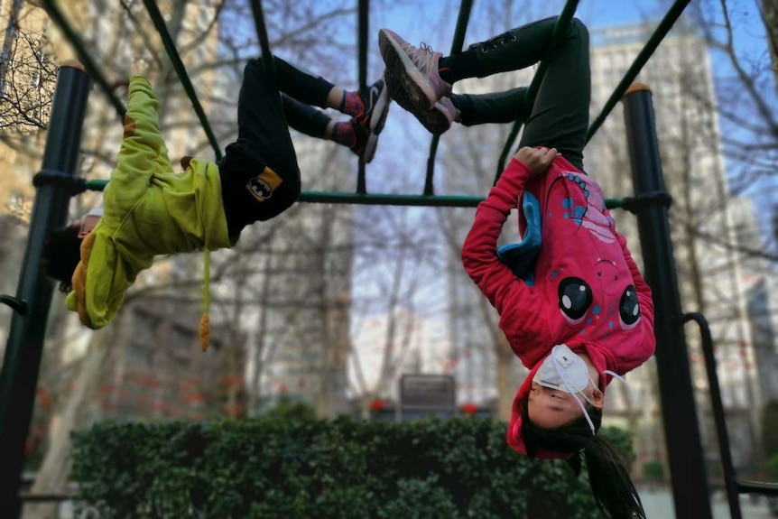 Two children wearing face masks hang upside down on a set of outdoor monkey bars.