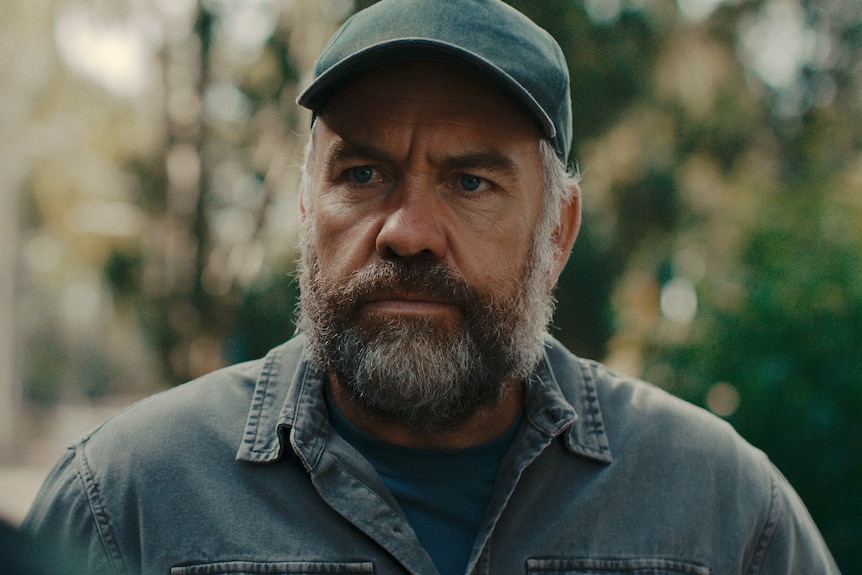 A TV still of Brendan Cowell, 50, bearded, in a cap, with an intense expression, brow furrowed.