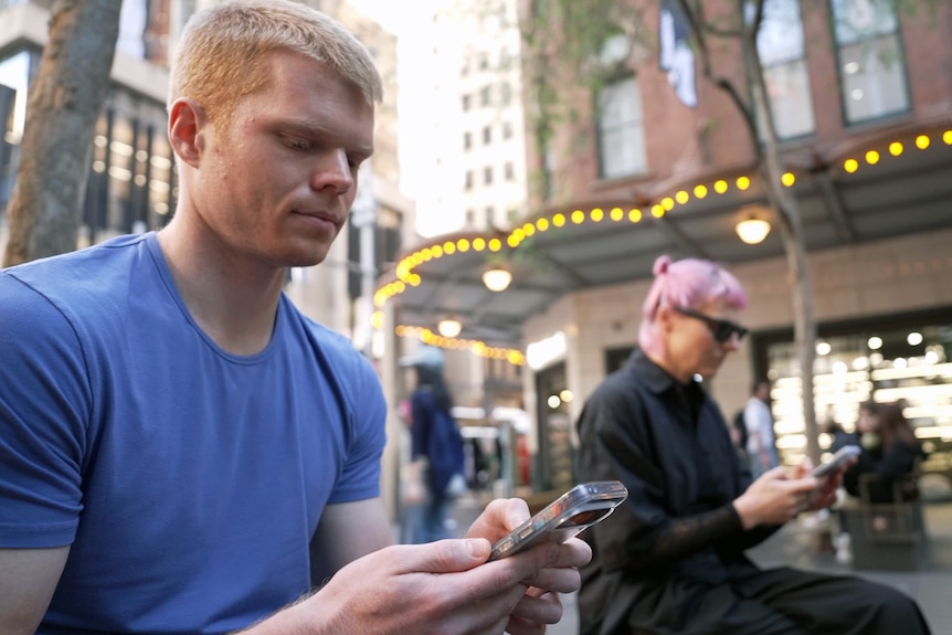 A man wearing a blue t-shirt sits on a public street holding a phone, a woman in the background wears black glasses.