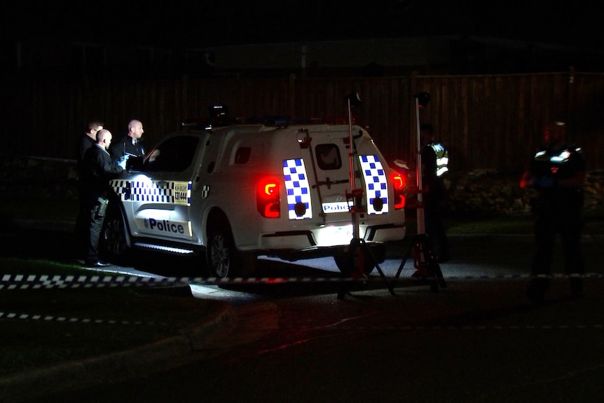 A number of police officers stand around a white and blue police vehicle at night.
