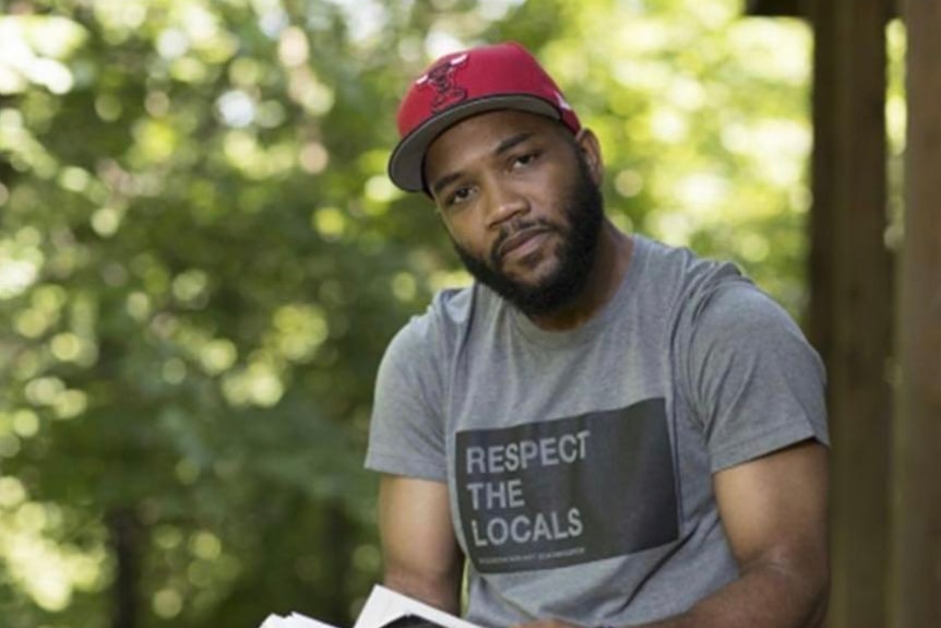 A Black man wearing a cap and holding a book, beard, moustache, his t-shirt has Respect the Locals written on it