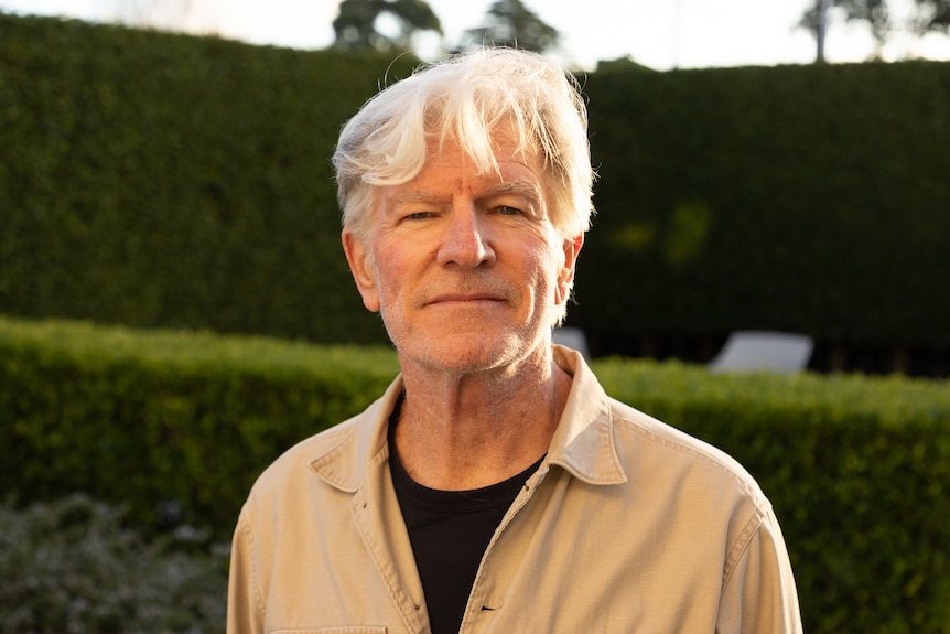Tim Finn with grey hair and a mustard shirt poses in front of a garden.