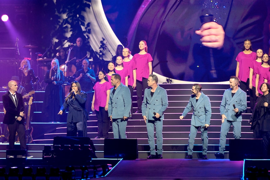 Performers on stage wearing grey suits; behind them are performers in pink shirts.