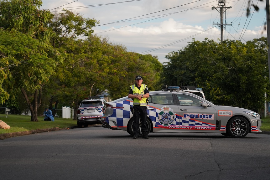 A police officer standing on a road wearing a yellow high-vis vest in front of a police car.