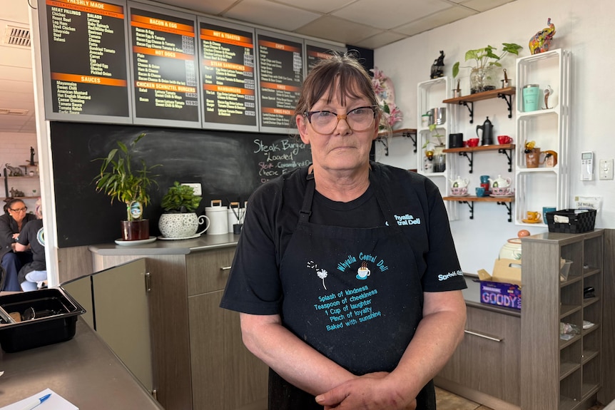 A woman wearing an apron and glasses is standing behind the counter in a cafe