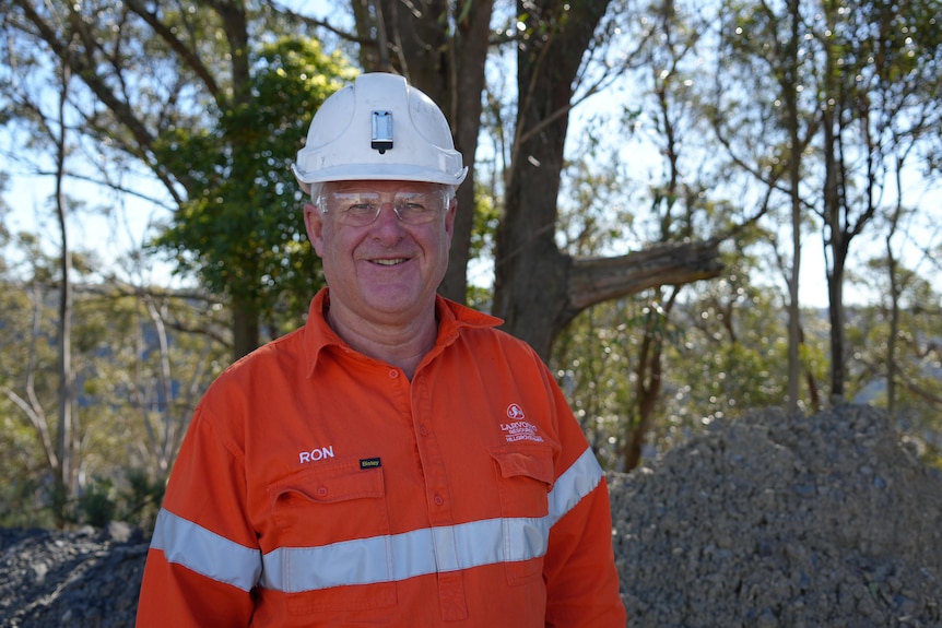 A man wearing high vis, hard hat and glasses smiles into camera with trees in the background 