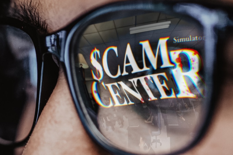 A man looks at the screen with the words Scam Center Simulator reflected in his glasses