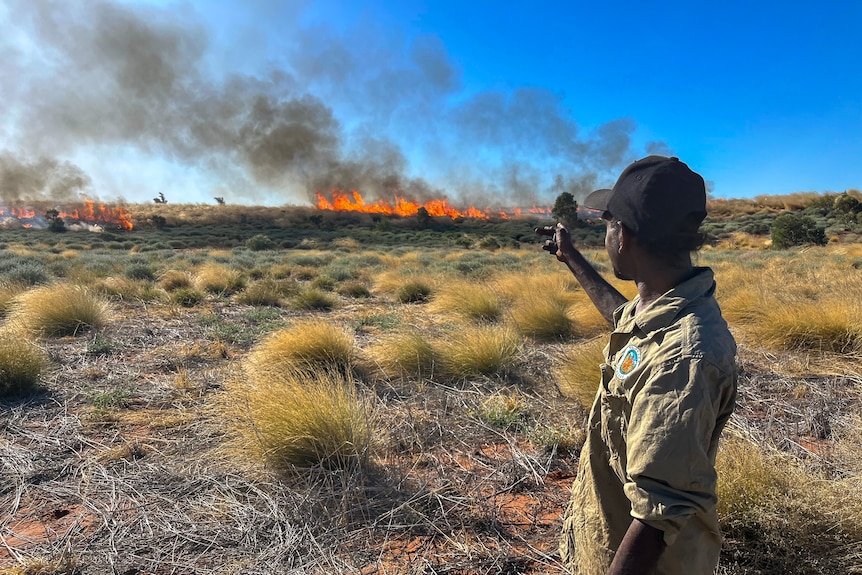 A man wearing a khaki shirt and black cap points to a fire burning in the distance.