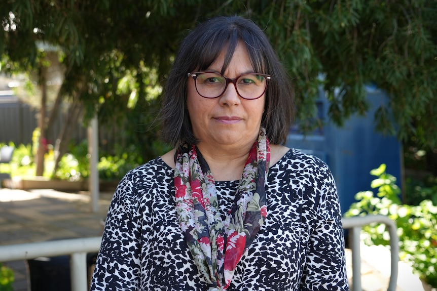 A woman with dark brown hair and glasses looks at the camera