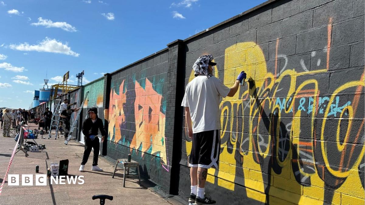 Artists paint murals on a large seaside wall. Some have ladders and stands. A man closest to the camera is spray painting yellow paint on to the black wall.