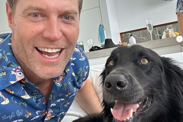 A smiling, fair-haired man with a dark-coloured dog in a house.