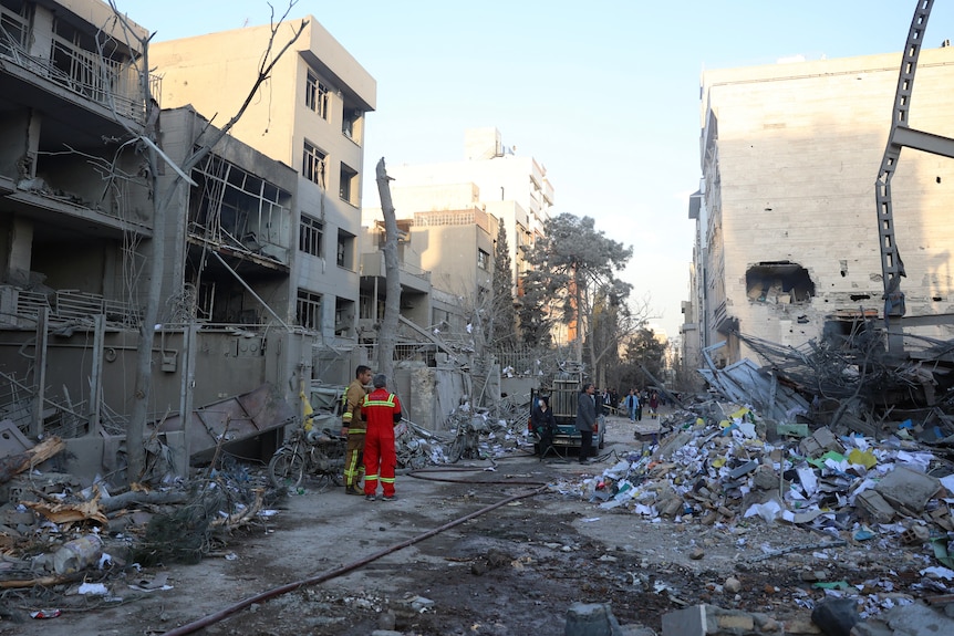 A pair of firefighters stand in a street surrounded by damaged buildings and rubble.