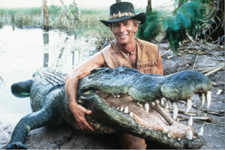 Paul Hogan dressed as Mick Dundee, holding a crocodile around the neck and smiling as he sits on bank of waterway.