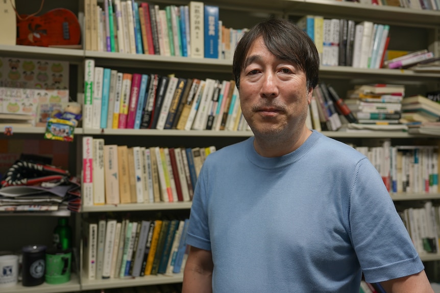 Ippei, a Japanese man wearing a blue t-shirt standing in front of shelves of books, slight smile.