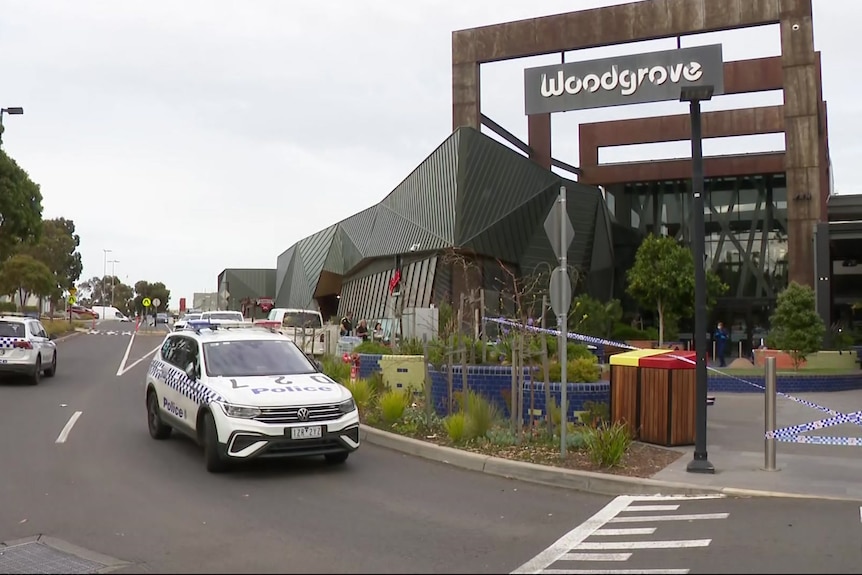 Two police cars are parked outside a shopping centre with a sign saying "Woodgrove" above police tape.