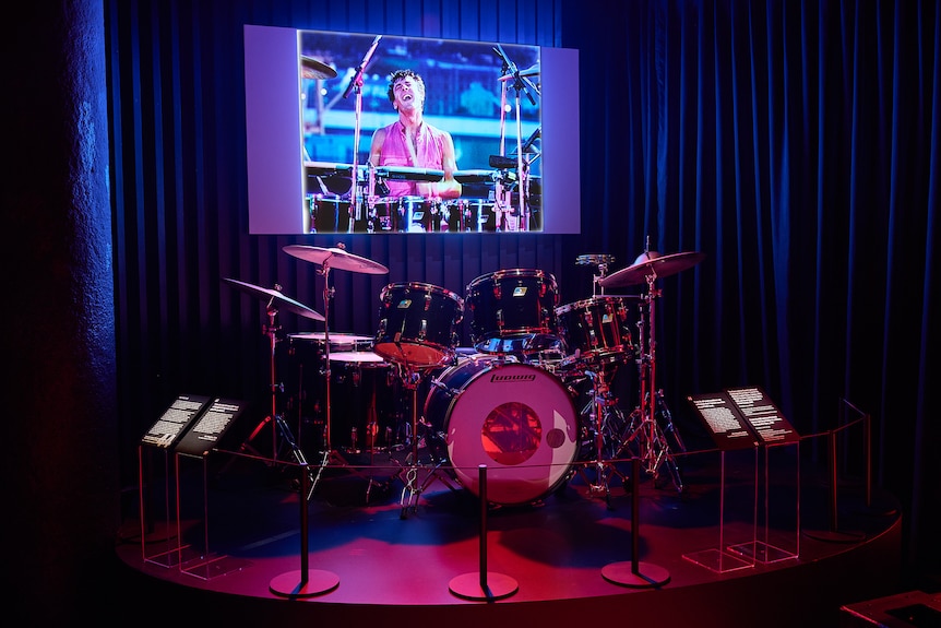 A drum kit sits on a slightly raised stage in a dark room with a screen behind showing a man playing the drums.