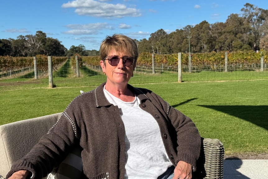 A woman with short hair wearing sunglasses and a brown cardigan relaxes on a chair at a vineyard.