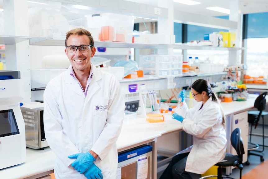 A man in a lab coat, gloves, and glasses smiling with a lab and lab tech in the background