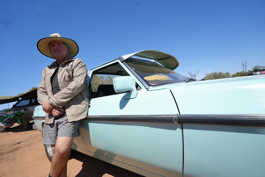 Man with wide-brim hat leans on old car and looks ahead 