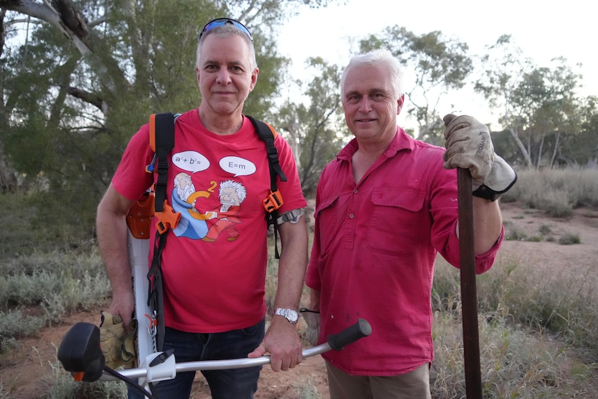 Ben McIntyre and Steve Eldridge pose for the camera holding gardening tools with buffel grass in the background.