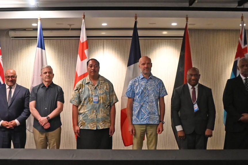 Five men stand in a line behind a desk, in front of a line of flags.
