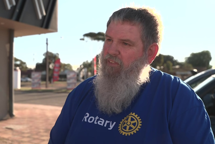 A man in a blue t-shirt and beard is standing on a footpath.