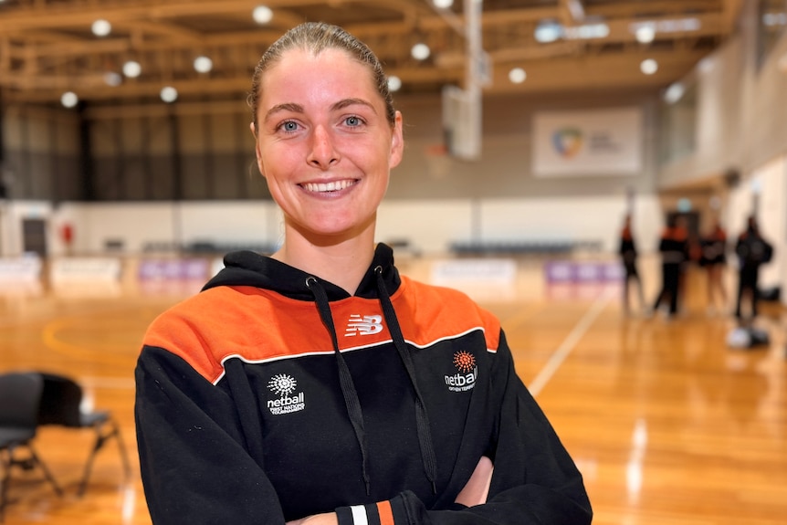 Netballer Gabby Coffey smiles for the camera, she wears a black and orange hoodie, with arms folded