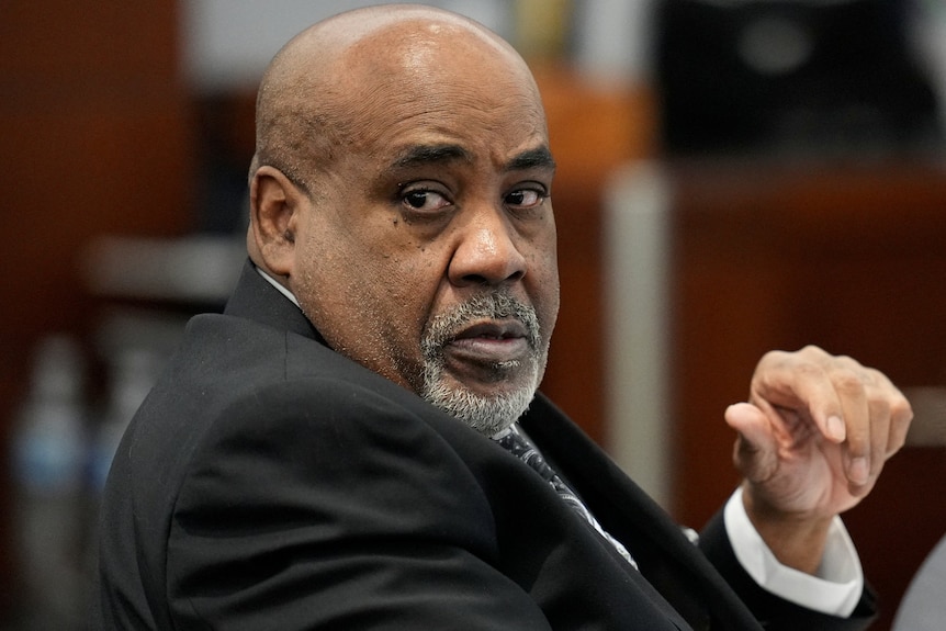 A heavy bald Black man with grey beard and moustache sitting in court wearing a suit, head turned towards the camera