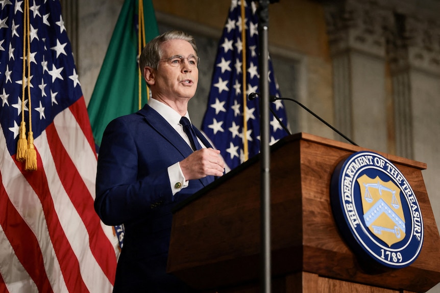 An older man with grey hair wearing a navy suit stands behind of a wooden lectern with US flags in the distance.