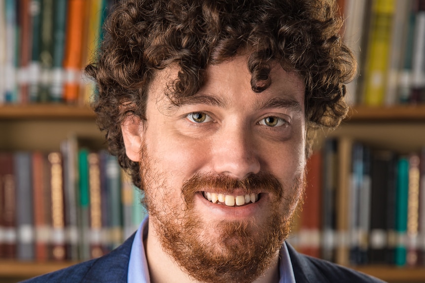 A close up of a smiling Tristan Buckmaster standing in front of a bookshelf