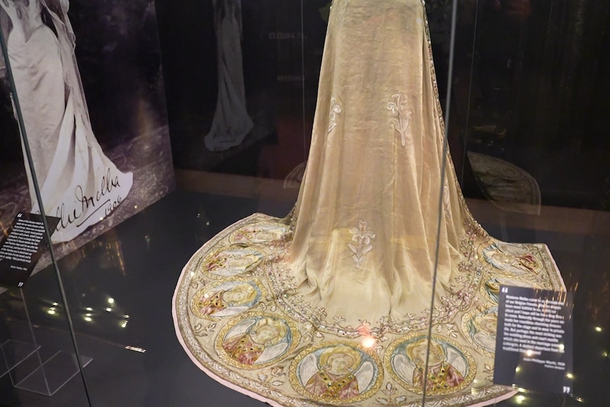 The bottom of a golden coloured cloak that is behind glass is spread out on the ground showing images of a woman.