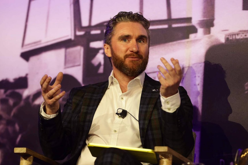 A man in a suit sits on a stage gesturing with his hands while talking