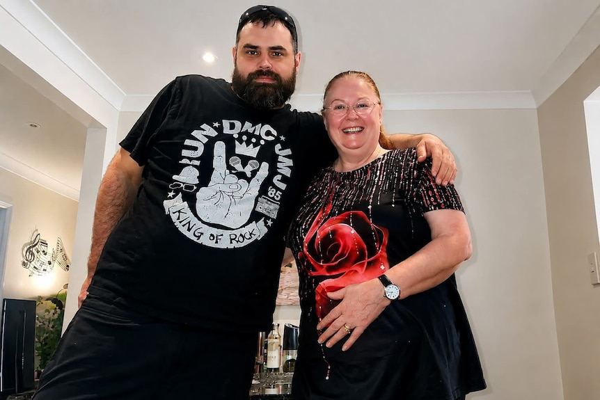 A dark-haired, bearded man stands with his arm around a smiling middle-aged woman in a living room.