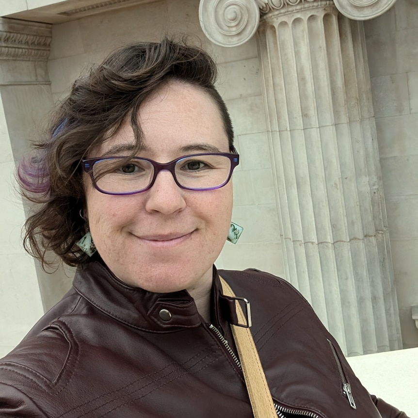 A woman with dark brown hair, glasses and a brown jacket in front a building with a classical-style pillar.