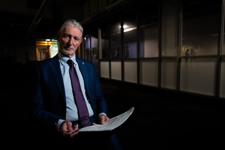 A man in a suit and tie sits in a darkened room clutching a piece of paper. 
