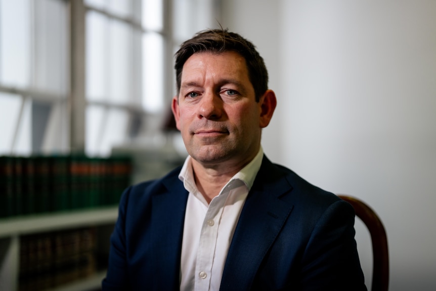 Close up of Luke Taylor, with brown hair, wearing a navy suit and white shirt smiles softly for photo near a book shelf.