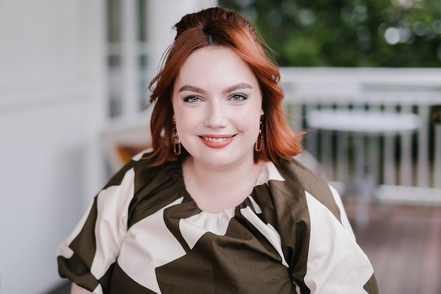 A woman with vivid red short hair wears a brown and cream print dress and smiles at the camera on a front porch