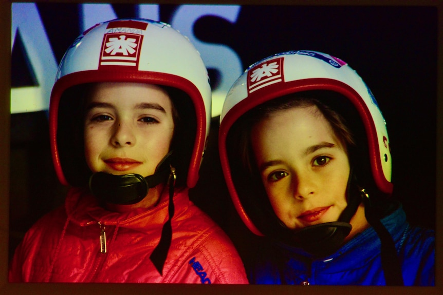 Two identical twins are next to each other, wearing helmets and smiling slightly at the camera.