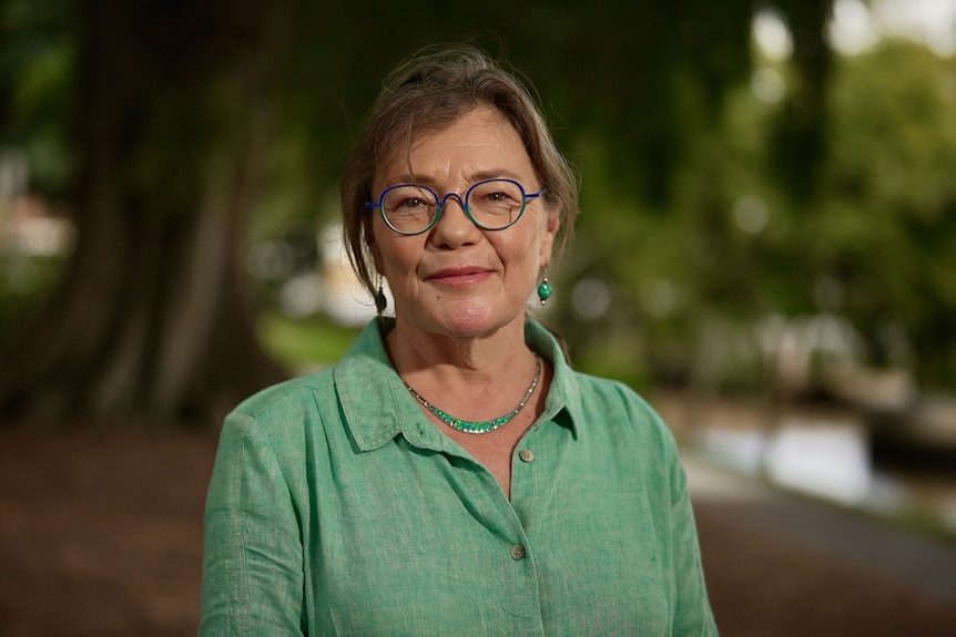 middle aged woman with green shirt, necklace, and glasses looking at camera, blurry river and tree behind her
