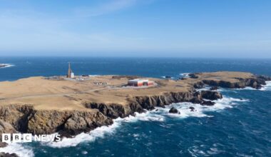 An aerial view of a long flat peninsula surrounded by dark blue sea. The coastline is rocky and the sky is blue. The island is brown and sparsely populated by several industrial buildings and a tall launch pad.