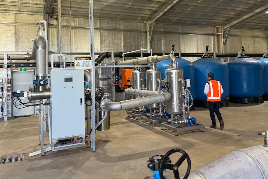 A metal system of pipes and cylinders stands in front of large storage tanks in a water processing facility