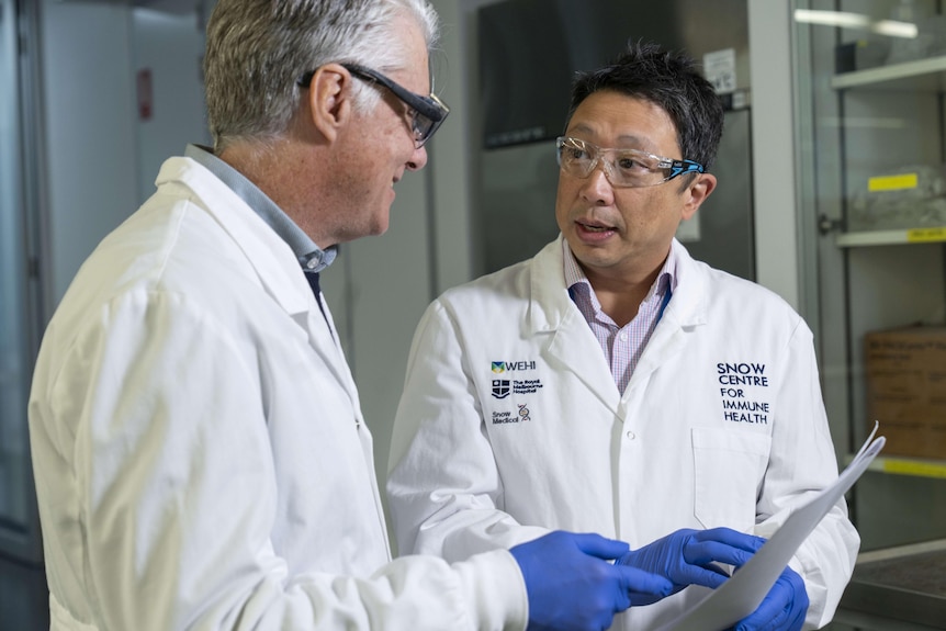 Two men in white lab coats look at a clipboard as they talk.