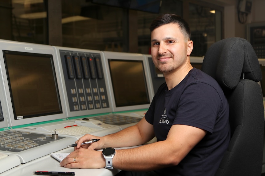 Nathan sits at a desk with screens and buttons, wearing a dark navy t-shirt.