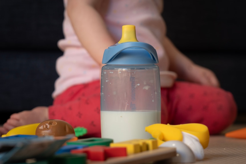 A baby bottle sitting on a table with children's toys and a baby sitting in the background.